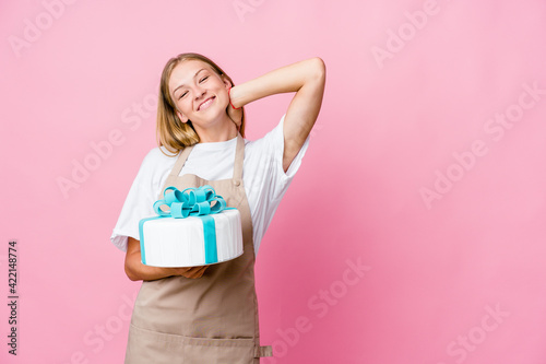 Young russian baker woman holding a delicious cake touching back of head, thinking and making a choice.