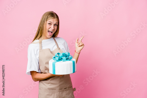 Young russian baker woman holding a delicious cake pointing with forefingers to a copy space, expressing excitement and desire.