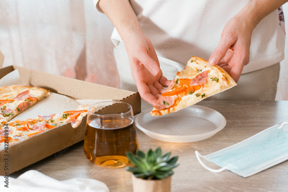 Takeout food. A woman puts a slice of pizza in a disposable plastic plate and a box of pizza on the table in the kitchen.
