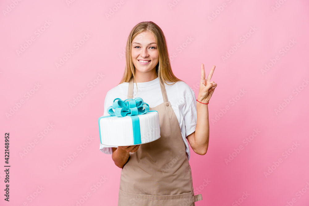 Young russian baker woman holding a delicious cake showing number two with fingers. Young russian baker woman holding a delicious cake showing number two with fingers.