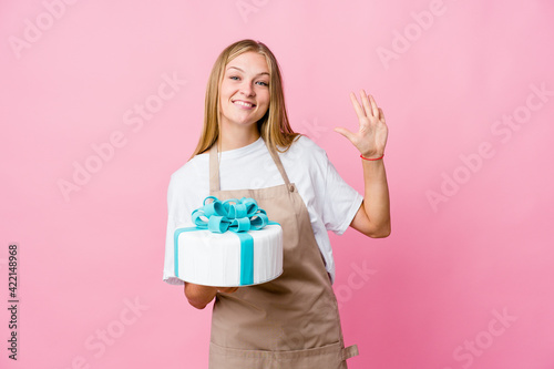 Young russian baker woman holding a delicious cake smiling cheerful showing number five with fingers.