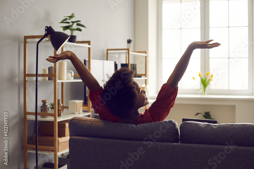 Young black lady enjoying weekend and relaxing on comfortable couch at home. African American woman sitting on sofa in living-room, stretching arms and smiling feeling untroubled and happy, back view