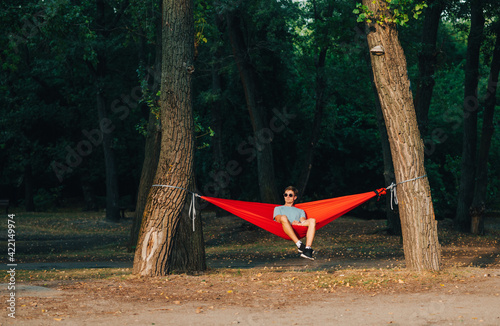 Young man in stylish casual clothes and sunglasses sitting in a hammock on a background of the park and admiring the sunset. Relax outdoors.