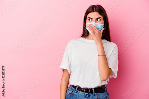 Young caucasian woman wearing a mask for virus isolated on pink background thoughtful looking to a copy space covering mouth with hand.