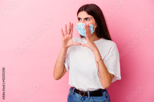 Young caucasian woman wearing a mask for virus isolated on pink background shouts loud, keeps eyes opened and hands tense.