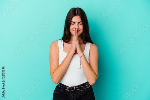 Young caucasian woman isolated on blue background holding hands in pray near mouth, feels confident. © Asier
