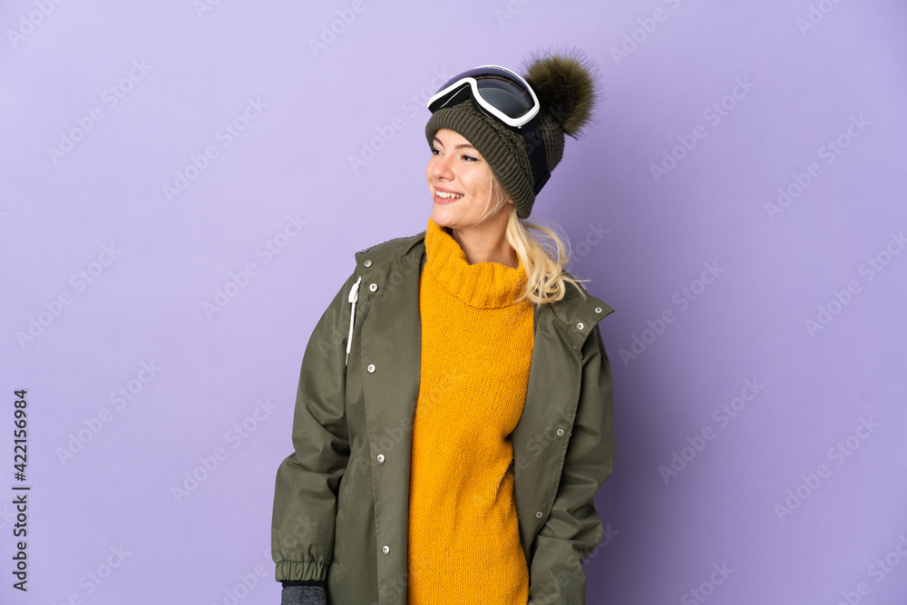 Skier Russian girl with snowboarding glasses isolated on purple background looking to the side and smiling