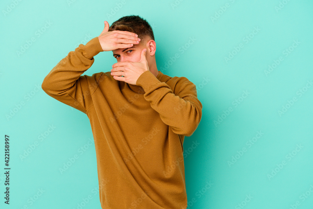 Young caucasian man isolated on blue background blink at the camera through fingers, embarrassed covering face. Young caucasian man isolated on blue background blink at the camera through fingers, embarrassed covering face.