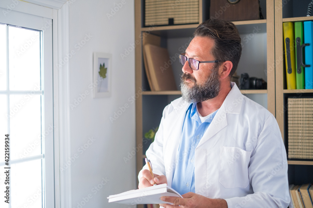 Fototapeta premium Pensive young male caucasian doctor in white medical uniform look in window distance thinking or pondering, serious man GP plan future career or success in medicine, visualize at workplace writing.