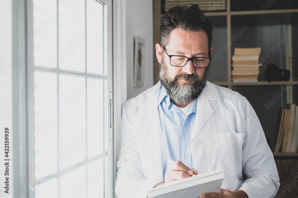 Fototapeta premium Pensive young male caucasian doctor in white medical uniform look in window distance thinking or pondering, serious man GP plan future career or success in medicine, visualize at workplace writing.