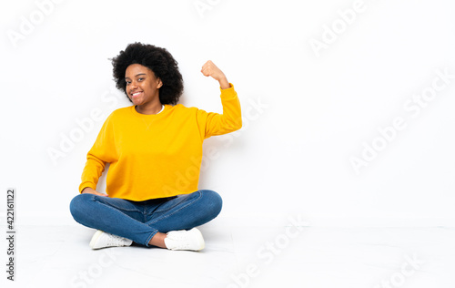 Young African American woman sitting on the floor doing strong gesture © luismolinero