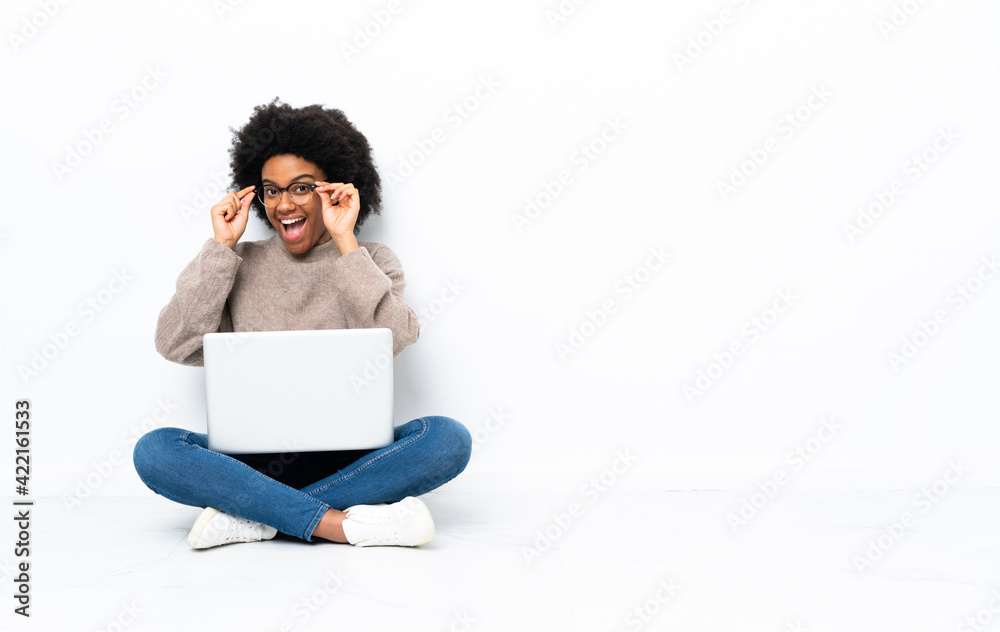 Young African American woman with a laptop sitting on the floor with glasses and surprised Young African American woman with a laptop sitting on the floor with glasses and surprised