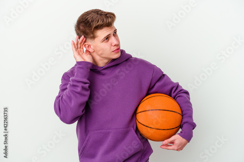 Young caucasian man playing basketball isolated background trying to listening a gossip.