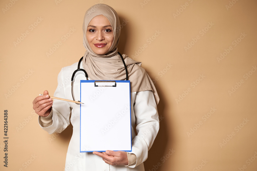 Charming female doctor in hijab pointing with a pencil at a blank sheet of paper on a clipboard. Healthcare and insurance concept. Copy space for promotion Charming female doctor in hijab pointing with a pencil at a blank sheet of paper on a clipboard. Healthcare and insurance concept. Copy space for promotion