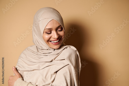 Adorable Muslim woman laughing with closed eyes on beige background with copy space © Taras Grebinets