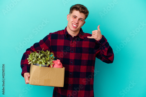 Young caucasian man moving holding a box isolated on blue background showing a mobile phone call gesture with fingers.