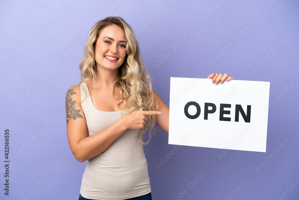 Young Brazilian woman isolated on purple background holding a placard with text OPEN and pointing it Young Brazilian woman isolated on purple background holding a placard with text OPEN and pointing it