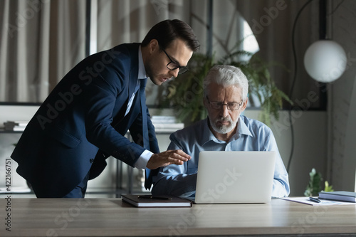 Young man CEO or boss help middle-aged colleague with computer in office. Diverse male coworkers brainstorm cooperate using laptop at workplace. Businessmen engaged in online work. Teamwork concept.
