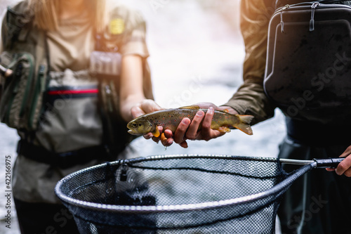 Young adult couple is fishing together on fast mountain river. They are holding a live trout before releasing it into the river again.