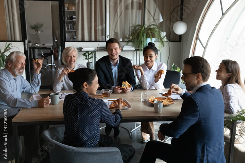 Smiling diverse employees sit in office have fun enjoy pizza on work lunch break together. Happy multiethnic colleagues coworkers laugh joke eating Italian fast food meal  drink coffee at workplace.
