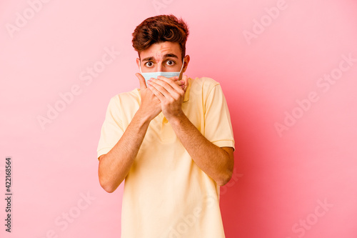 Young caucasian man wearing a protection for coronavirus isolated on pink background shocked covering mouth with hands.
