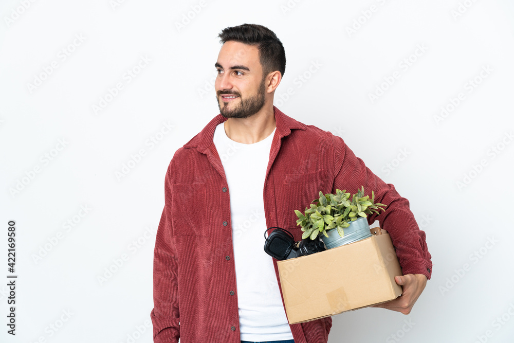 Young caucasian man making a move while picking up a box full of things isolated on white background looking to the side and smiling Young caucasian man making a move while picking up a box full of things isolated on white background looking to the side and smiling