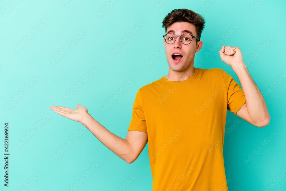 Young caucasian man isolated on blue background holds copy space on a palm, keep hand over cheek. Amazed and delighted. Young caucasian man isolated on blue background holds copy space on a palm, keep hand over cheek. Amazed and delighted.