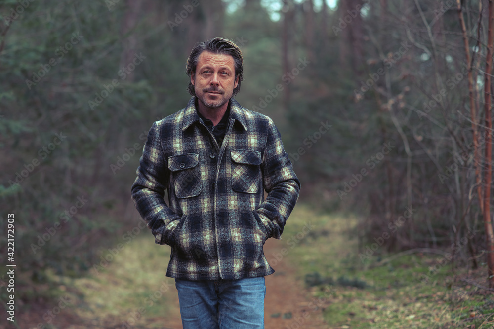 Cheerful blonde man in checkered coat walking on a path in a forest.