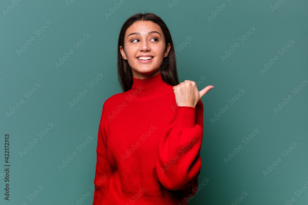Young Indian woman isolated on blue background points with thumb finger away, laughing and carefree.