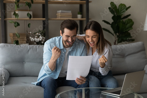 Overjoyed young couple reading good news in letter together, excited wife and husband celebrating success, showing yes gesture, approved loan or mortgage, money refund, sitting on couch at home