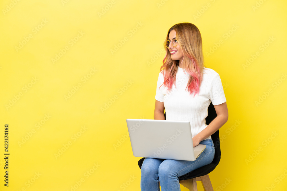 Fototapeta premium Young woman sitting on a chair with laptop over isolated yellow background looking side