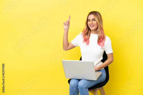 Young woman sitting on a chair with laptop over isolated yellow background pointing up a great idea