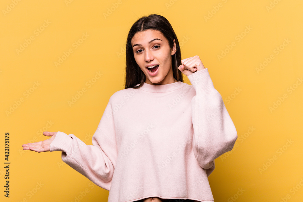 Young Indian woman isolated on yellow background holds copy space on a palm, keep hand over cheek. Amazed and delighted. Young Indian woman isolated on yellow background holds copy space on a palm, keep hand over cheek. Amazed and delighted.