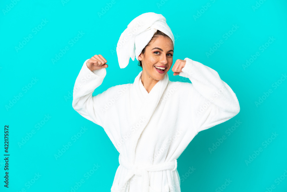 Young woman in a bathrobe over isolated blue background doing strong gesture Young woman in a bathrobe over isolated blue background doing strong gesture