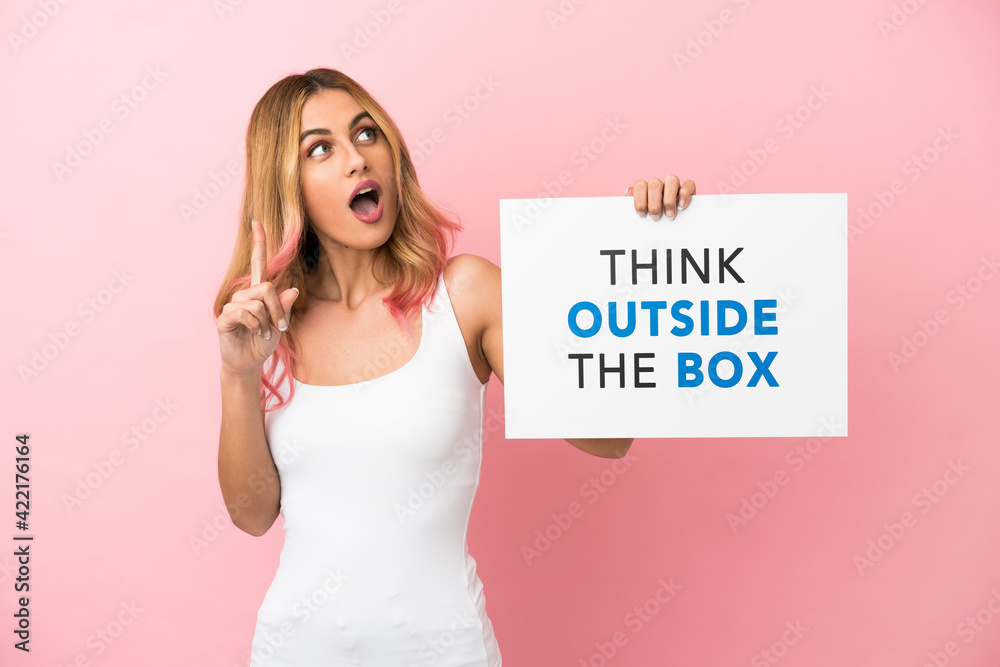 Young woman over isolated pink background holding a placard with text Think Outside The Box and thinking Young woman over isolated pink background holding a placard with text Think Outside The Box and thinking