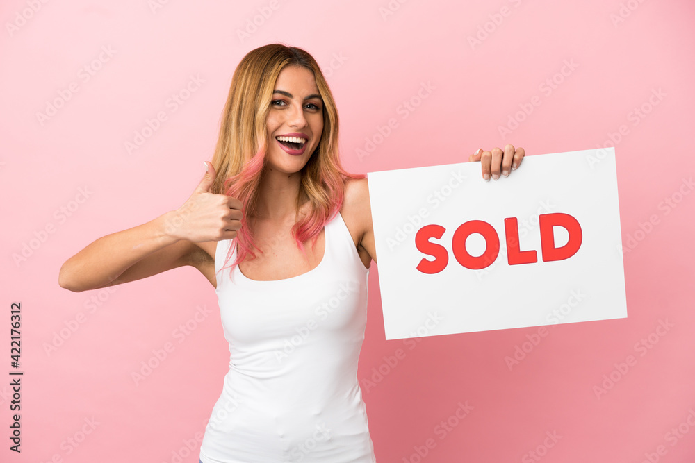 Young woman over isolated pink background holding a placard with text SOLD with thumb up Young woman over isolated pink background holding a placard with text SOLD with thumb up
