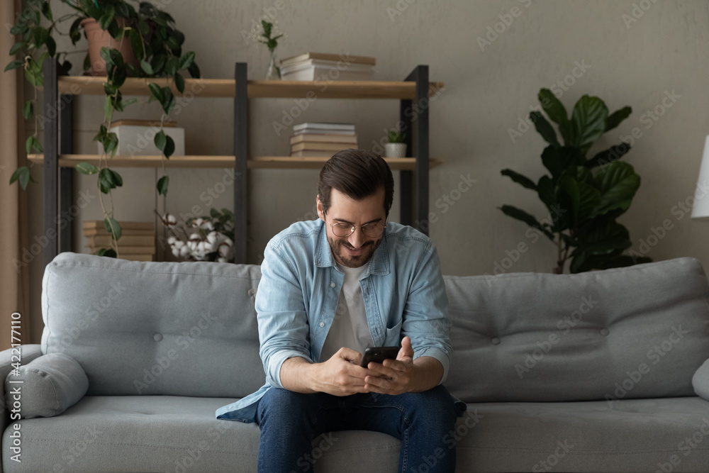 Smiling man wearing glasses having fun with phone at home, sitting on couch, looking at mobile device screen, chatting or shopping online, browsing apps, enjoying leisure time with smartphone