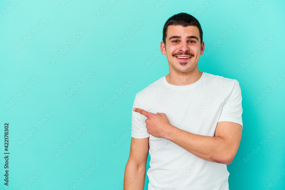 Obraz premium Young caucasian man isolated on blue background smiling and pointing aside, showing something at blank space.