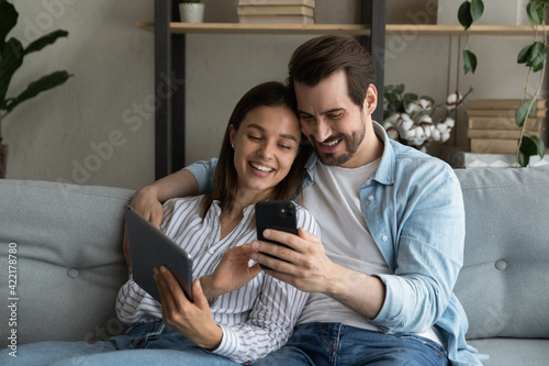 Close up happy young couple having fun with mobile devices together, using smartphone and tablet, smiling woman and man hugging, looking at phone screen, chatting or shopping online at home