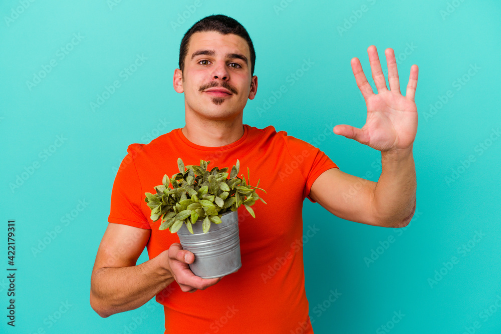 Young caucasian man holding a leaf isolated on blue background smiling cheerful showing number five with fingers. Young caucasian man holding a leaf isolated on blue background smiling cheerful showing number five with fingers.