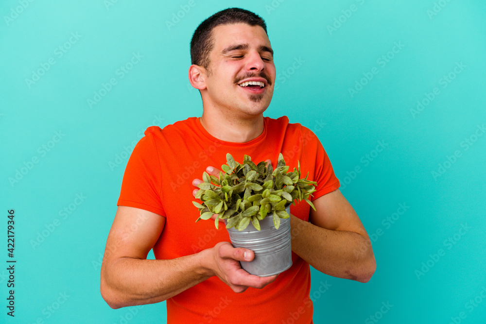 Young caucasian man holding a leaf isolated on blue background laughs out loudly keeping hand on chest. Young caucasian man holding a leaf isolated on blue background laughs out loudly keeping hand on chest.
