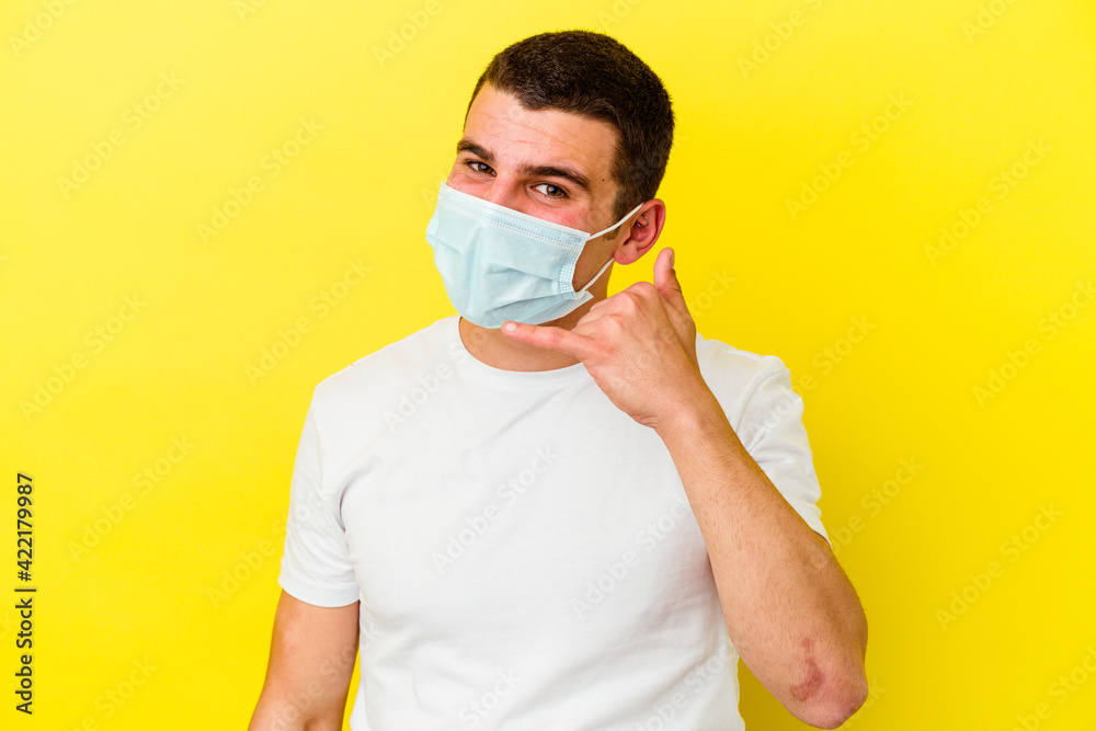 Young caucasian man wearing a protection for coronavirus isolated on yellow background showing a mobile phone call gesture with fingers. Young caucasian man wearing a protection for coronavirus isolated on yellow background showing a mobile phone call gesture with fingers.