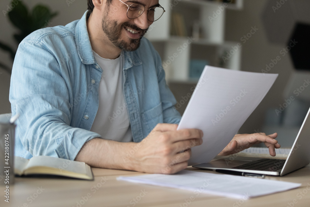 Close up smiling businessman in glasses using laptop, holding paper documents, typing, happy man consulting client online or working on financial report, student searching information for project Close up smiling businessman in glasses using laptop, holding paper documents, typing, happy man consulting client online or working on financial report, student searching information for project