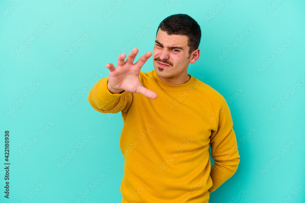 Young caucasian man isolated on blue background showing claws imitating a cat, aggressive gesture.