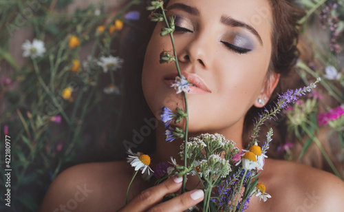 Beauty close up portrait of pretty girl lying on wildflowers with flowers around her head. View from above. Cute smiling young woman closed eyes. Concept of spring summer youth beauty. Beautiful face.