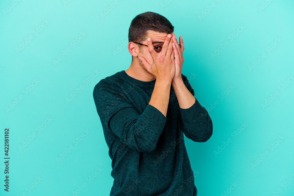 Young caucasian man isolated on blue background blink through fingers frightened and nervous. Young caucasian man isolated on blue background blink through fingers frightened and nervous.