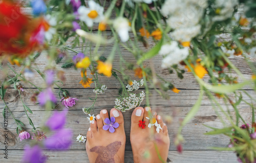 Summer lifestyle portrait of a attractive girl's feet with buds of wildflowers between toes. Staying on wooden terrace surrounded by flowers. Enjoying life, nature. Feet in focus, flowers in blur. 