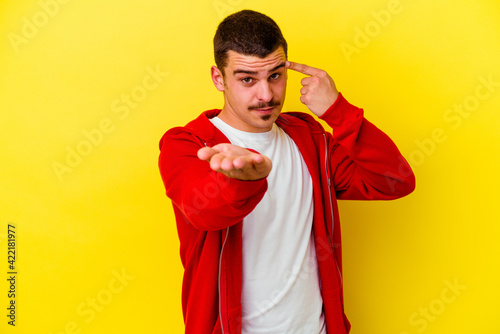 Young caucasian cool man isolated on yellow background holding and showing a product on hand.