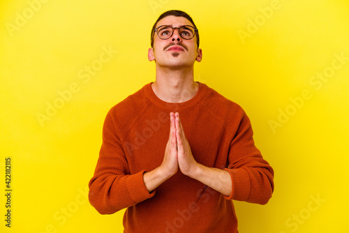 Young caucasian cool man isolated on yellow background holding hands in pray near mouth, feels confident.