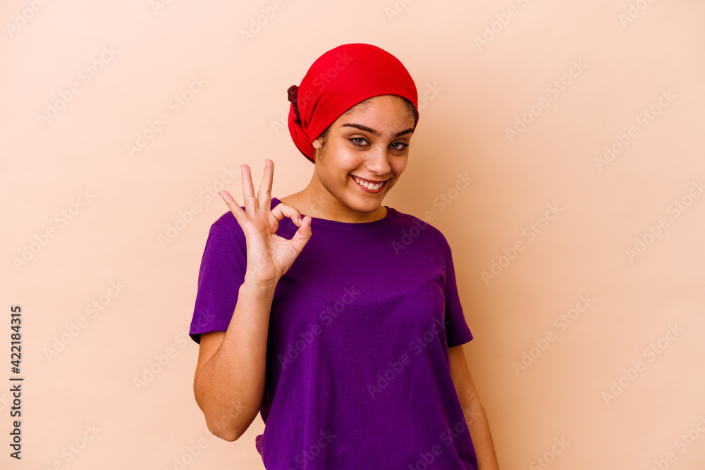 Young african american woman isolated on beige background winks an eye and holds an okay gesture with hand. Young african american woman isolated on beige background winks an eye and holds an okay gesture with hand.